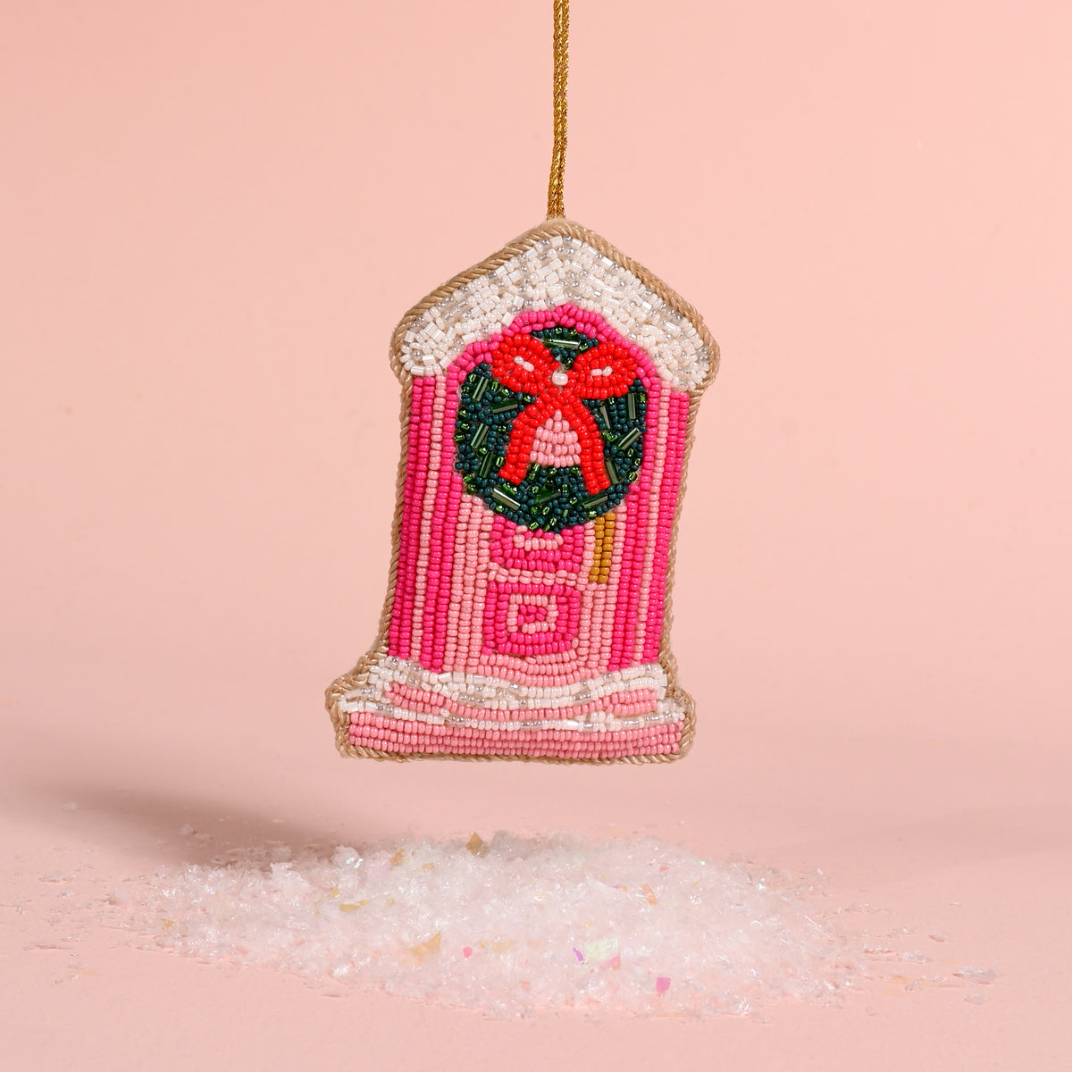 Darling Doorstep Handcrafted Beaded Ornament shaped like a pink front door, decorated with a festive wreath and red bow, hanging above a pile of artificial snow.