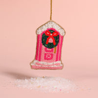 Darling Doorstep Handcrafted Beaded Ornament shaped like a pink front door, decorated with a festive wreath and red bow, hanging above a pile of artificial snow.