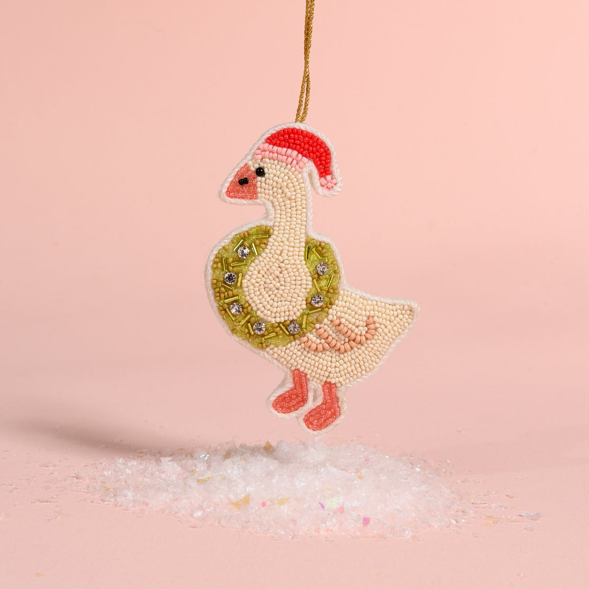 A Merry Silly Goose Handcrafted Beaded Ornament wearing a Santa hat and a green wreath around its neck, hanging above a small pile of artificial snow.