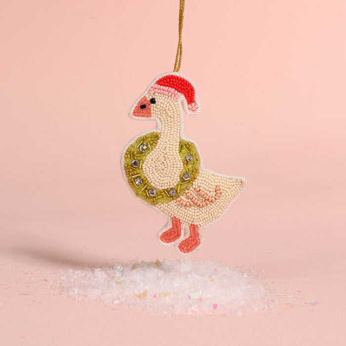 A Merry Silly Goose Handcrafted Beaded Ornament wearing a Santa hat and a green wreath around its neck, hanging above a small pile of artificial snow.
