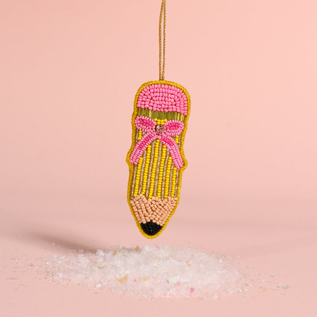 Pencil Handcrafted Beaded Ornament shaped like a pencil with a pink eraser and bow, hanging above a small pile of faux snow.