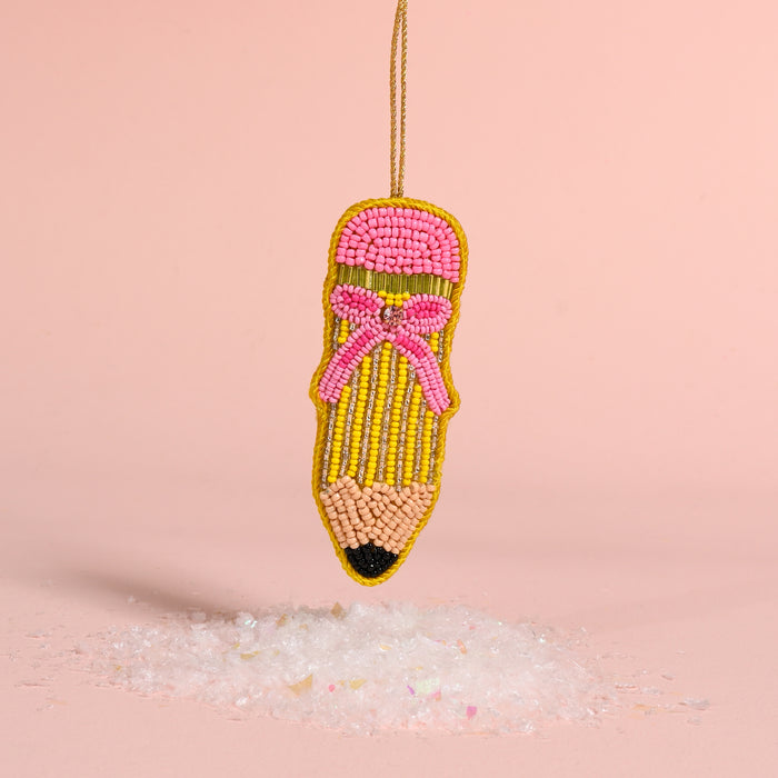 Pencil Handcrafted Beaded Ornament shaped like a pencil with a pink eraser and bow, hanging above a small pile of faux snow.