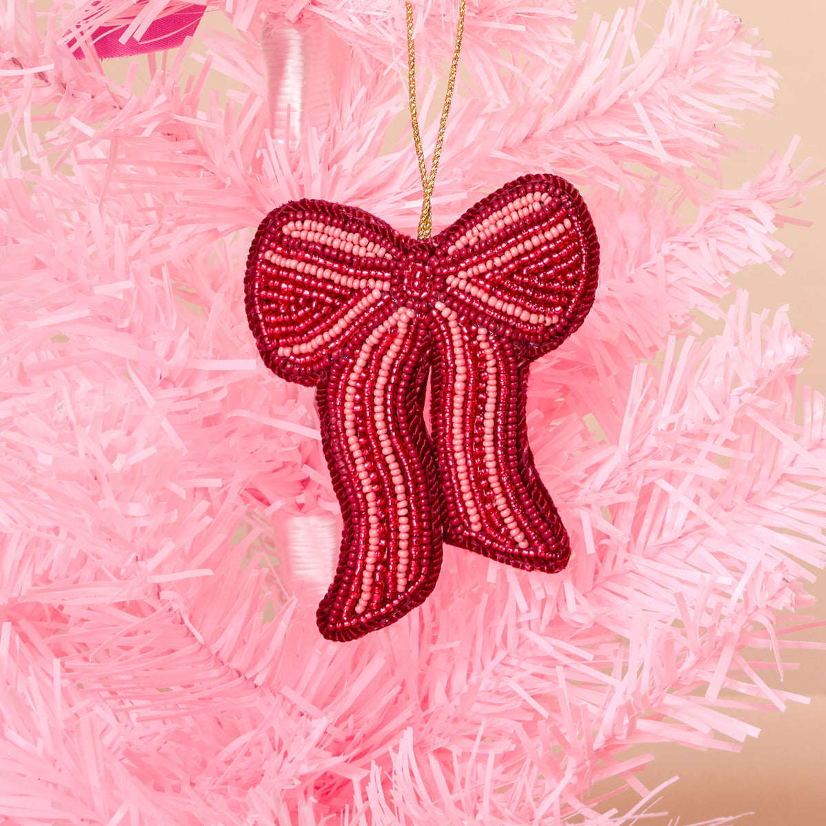 A Red Bow Handcrafted Beaded Ornament from The Darling Effect hangs on a pink artificial tree, adding a festive touch to the holiday décor.