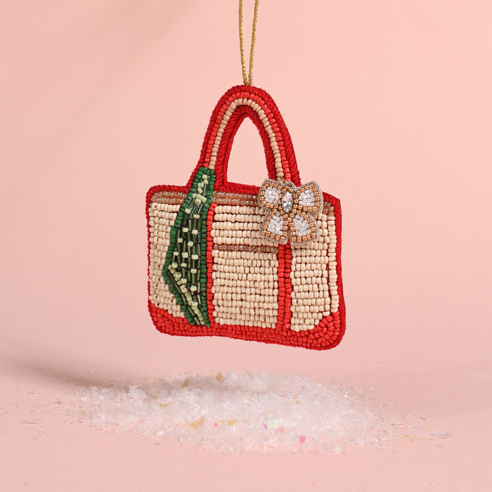 Tote of the Year Handcrafted Beaded Ornament shaped like a tote bag with red trim, a green striped accent, and a jeweled floral embellishment, hanging above a layer of faux snow.