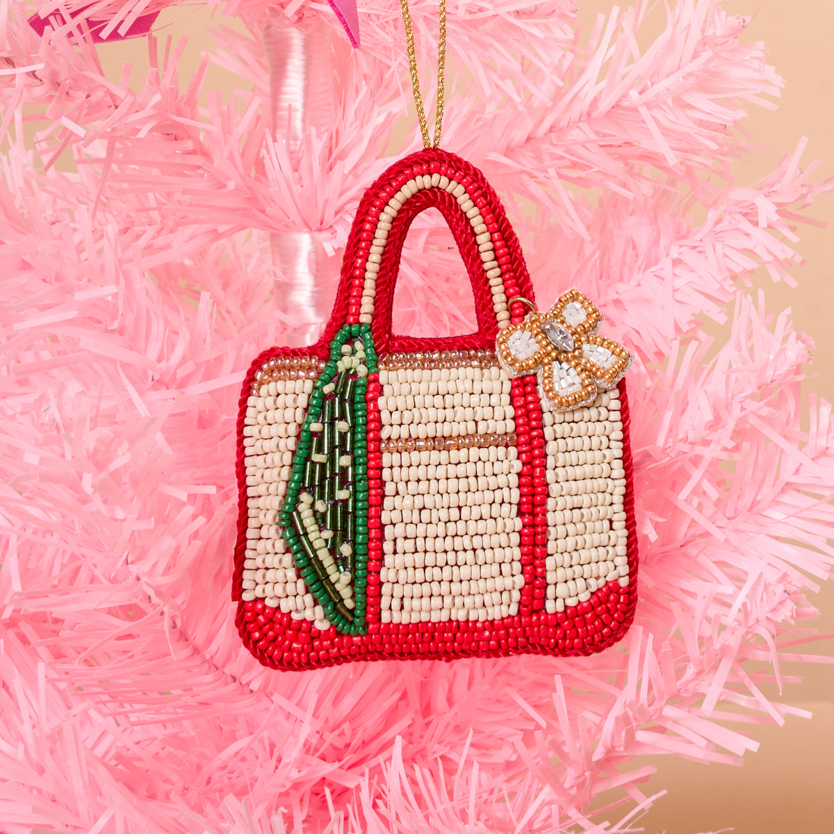 The Darling Effect Tote of the Year Handcrafted Beaded Ornament shaped like a tote bag with red and cream detailing, a green ribbon accent, and a bow charm, hanging on a pink Christmas tree.