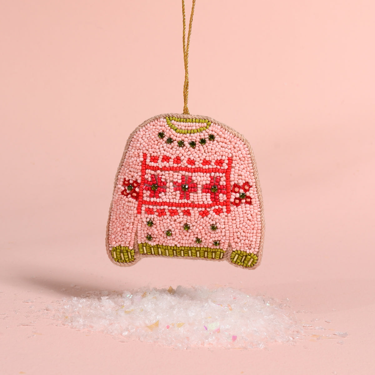 The Darling Effect Christmas Sweater Handcrafted Beaded Ornament, shaped like a pink sweater with red and green patterned details, hangs above a pile of faux snow as festive holiday décor.