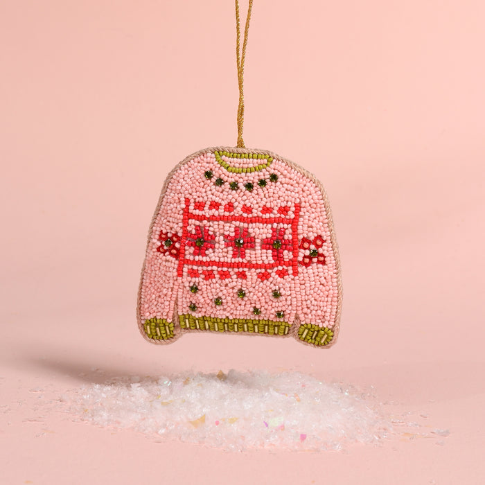 The Darling Effect Christmas Sweater Handcrafted Beaded Ornament, shaped like a pink sweater with red and green patterned details, hangs above a pile of faux snow as festive holiday décor.