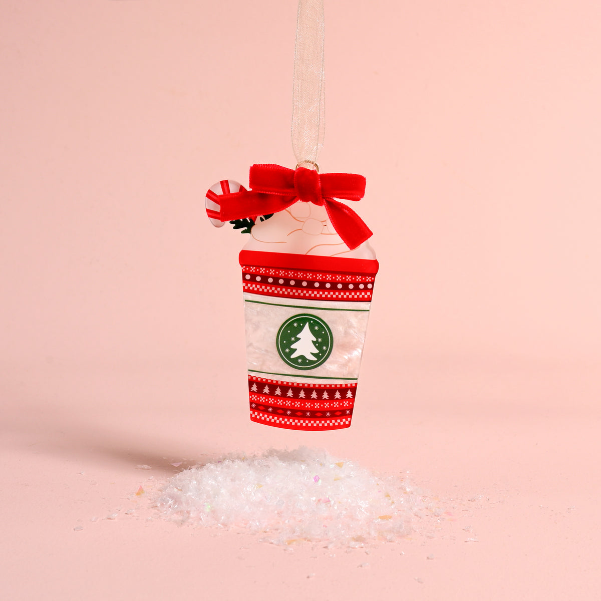 A Peppermint Latte Acrylic Ornament by The Darling Effect featuring a red bow, peppermint candy, and green tree logo, hanging above a small pile of artificial snow.