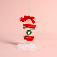 A Peppermint Latte Acrylic Ornament by The Darling Effect featuring a red bow, peppermint candy, and green tree logo, hanging above a small pile of artificial snow.