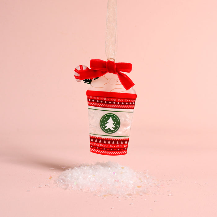 A Peppermint Latte Acrylic Ornament by The Darling Effect featuring a red bow, peppermint candy, and green tree logo, hanging above a small pile of artificial snow.