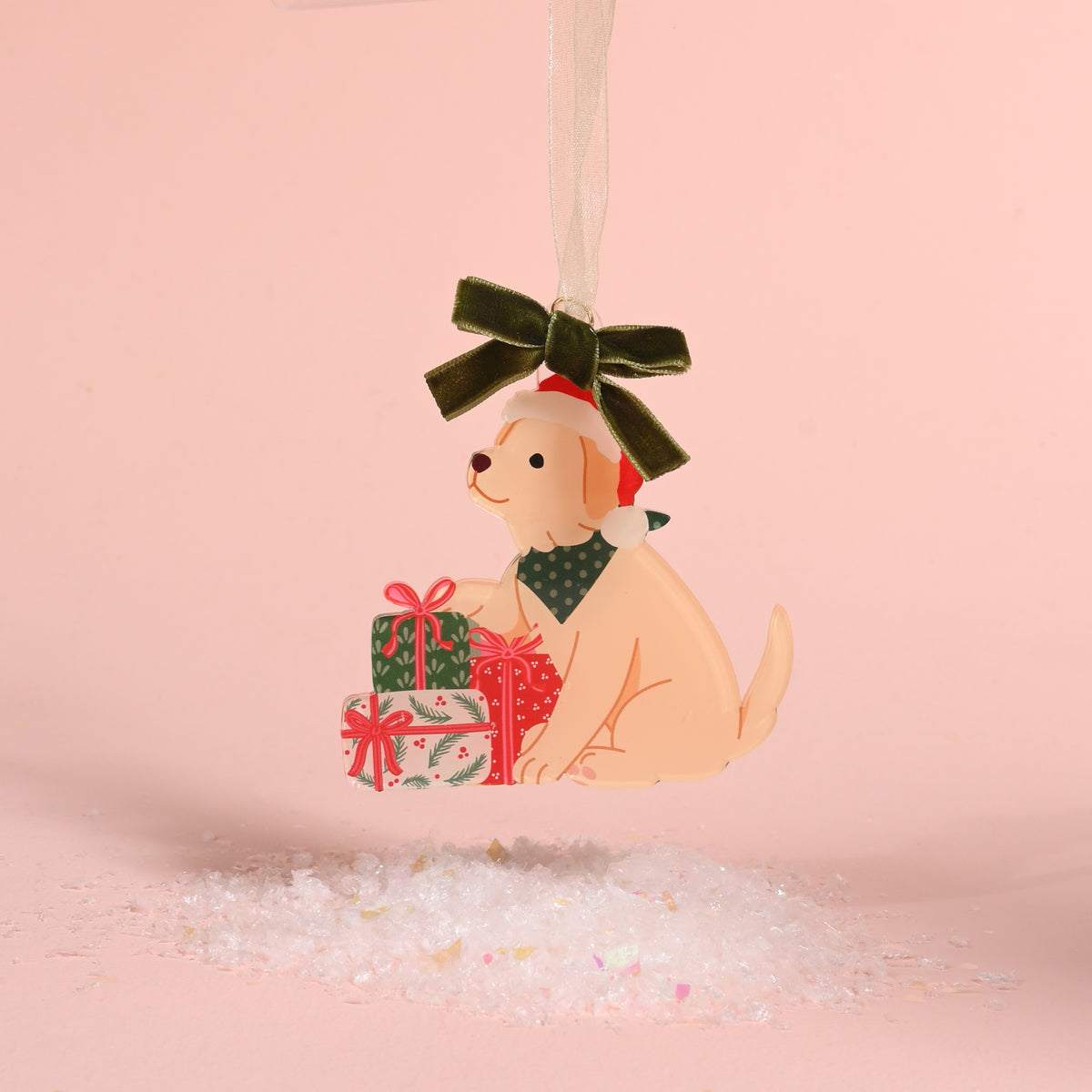 The Santa Paws Acrylic Ornament by The Darling Effect features a light-colored dog in a Santa hat and green bandana sitting by Christmas gifts, hanging from a green velvet ribbon.