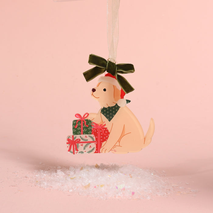 The Santa Paws Acrylic Ornament by The Darling Effect features a light-colored dog in a Santa hat and green bandana sitting by Christmas gifts, hanging from a green velvet ribbon.