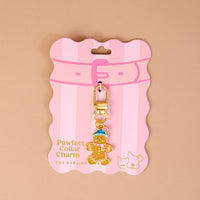 The Gingerbread Pawfect Collar Charm features a gingerbread man wearing a blue hat and pink striped scarf, attached to a gold lobster clasp and displayed on a pink striped backing card for pet accessories.