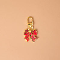The Holiday Bow Pawfect Collar Charm by The Darling Effect is a gold-toned metal pet accessory in the shape of a red bow, featuring a lobster clasp for easy attachment to collar charms.