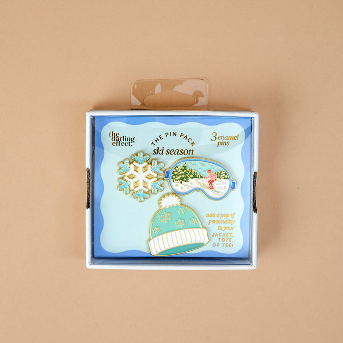 Ski Season The Pin Pack featuring three enamel pins: a snowflake, a snowy landscape inside ski goggles, and a winter beanie displayed in a box.