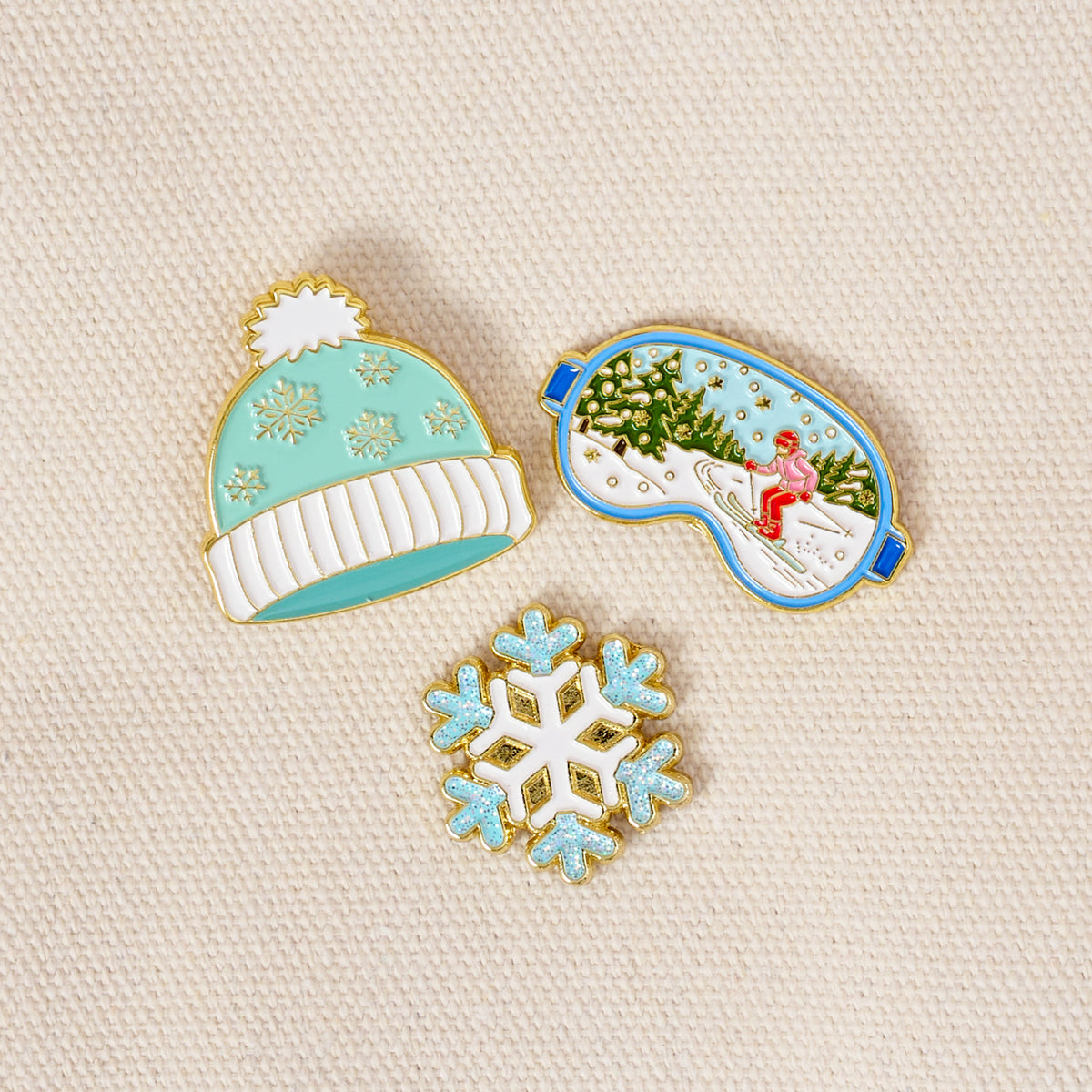 Ski Season The Pin Pack includes a mint beanie with a white pom-pom, a ski mask depicting a person skiing in a snowy forest, and a light blue snowflake.