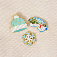 Ski Season The Pin Pack includes a mint beanie with a white pom-pom, a ski mask depicting a person skiing in a snowy forest, and a light blue snowflake.