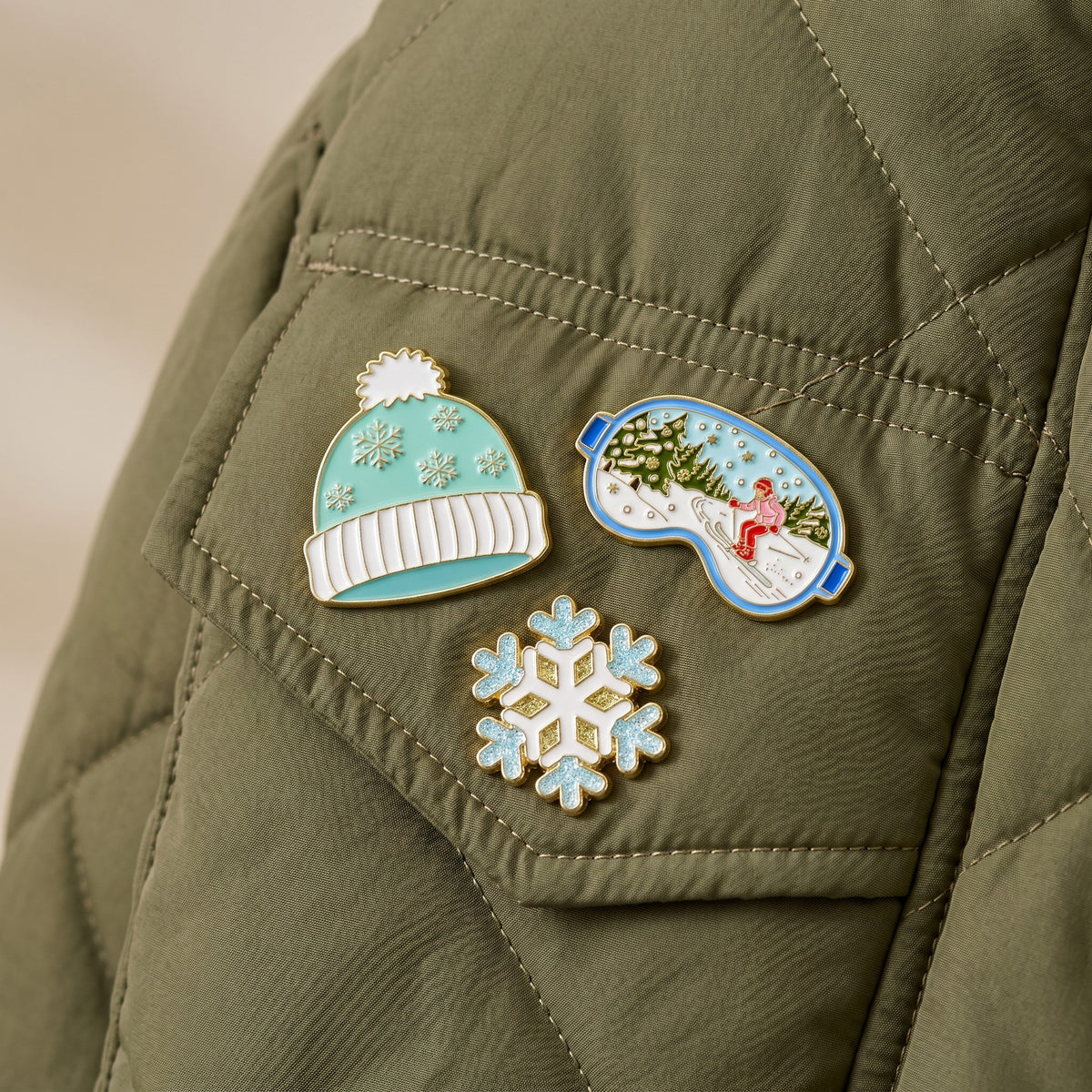 Three winter-themed enamel pins from The Darling Effect Ski Season The Pin Pack—a teal beanie, a snowflake, and ski goggles—pinned to a green quilted jacket.