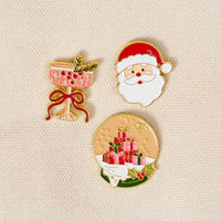 Christmas Cheer The Pin Pack featuring three enamel pins: a pink cocktail glass with a sprig, a Santa Claus face, and a snow globe containing a stack of wrapped gifts.