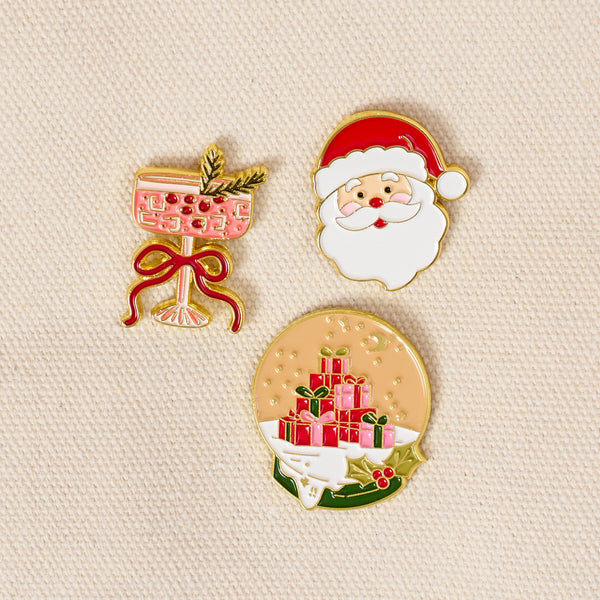 Christmas Cheer The Pin Pack featuring three enamel pins: a pink cocktail glass with a sprig, a Santa Claus face, and a snow globe containing a stack of wrapped gifts.