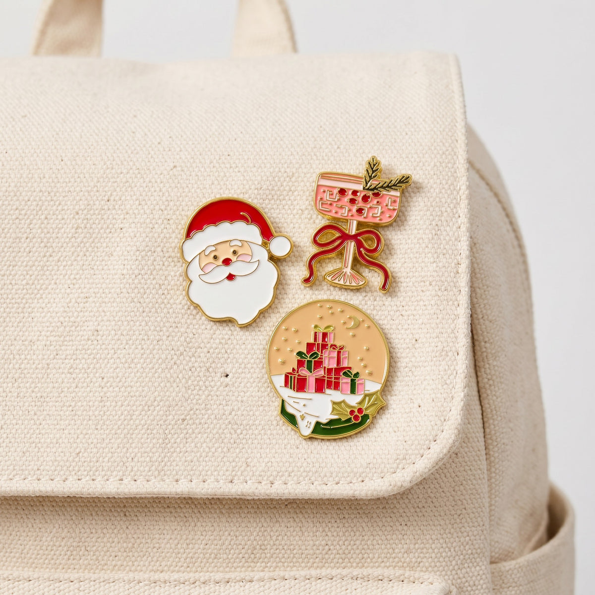 The Christmas Cheer The Pin Pack features three expressive enamel pins—a Santa face, a cocktail glass with a bow, and a snow globe containing a stack of presents—pinned to a canvas backpack.