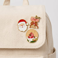 The Christmas Cheer The Pin Pack features three expressive enamel pins—a Santa face, a cocktail glass with a bow, and a snow globe containing a stack of presents—pinned to a canvas backpack.