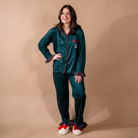 A woman wears The Darling Effect Evergreen Plaid Sweet Dreams Pajama Pants Set, featuring a dark green long-sleeved button-down top with red plaid trim and ruffles, paired with cream-colored slippers with red bows.