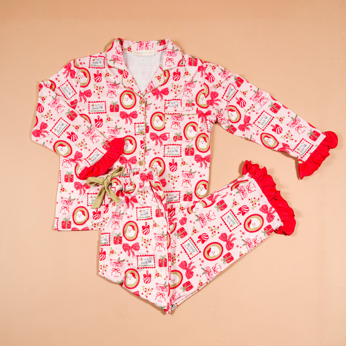The Darling Effect Mistletoe Magic Sweet Dreams Pajama Pants Set features a long-sleeved button-up top and matching pants in a pink and red Christmas-themed print, detailed with red ruffles at the wrists and pant hems.
