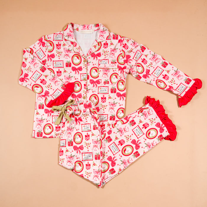The Darling Effect Mistletoe Magic Sweet Dreams Pajama Pants Set features a long-sleeved button-up top and matching pants in a pink and red Christmas-themed print, detailed with red ruffles at the wrists and pant hems.