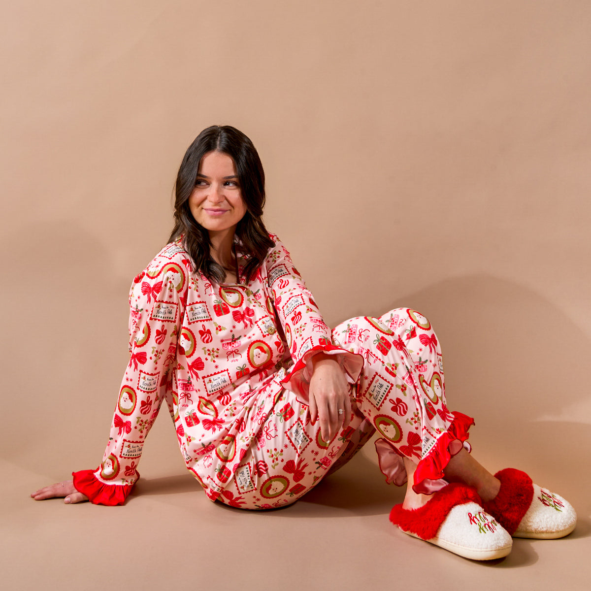 A woman sits on the floor wearing a red, Christmas-themed Mistletoe Magic Sweet Dreams Pajama Pants Set by The Darling Effect, featuring ruffled cuffs and matching fur-trimmed slippers.