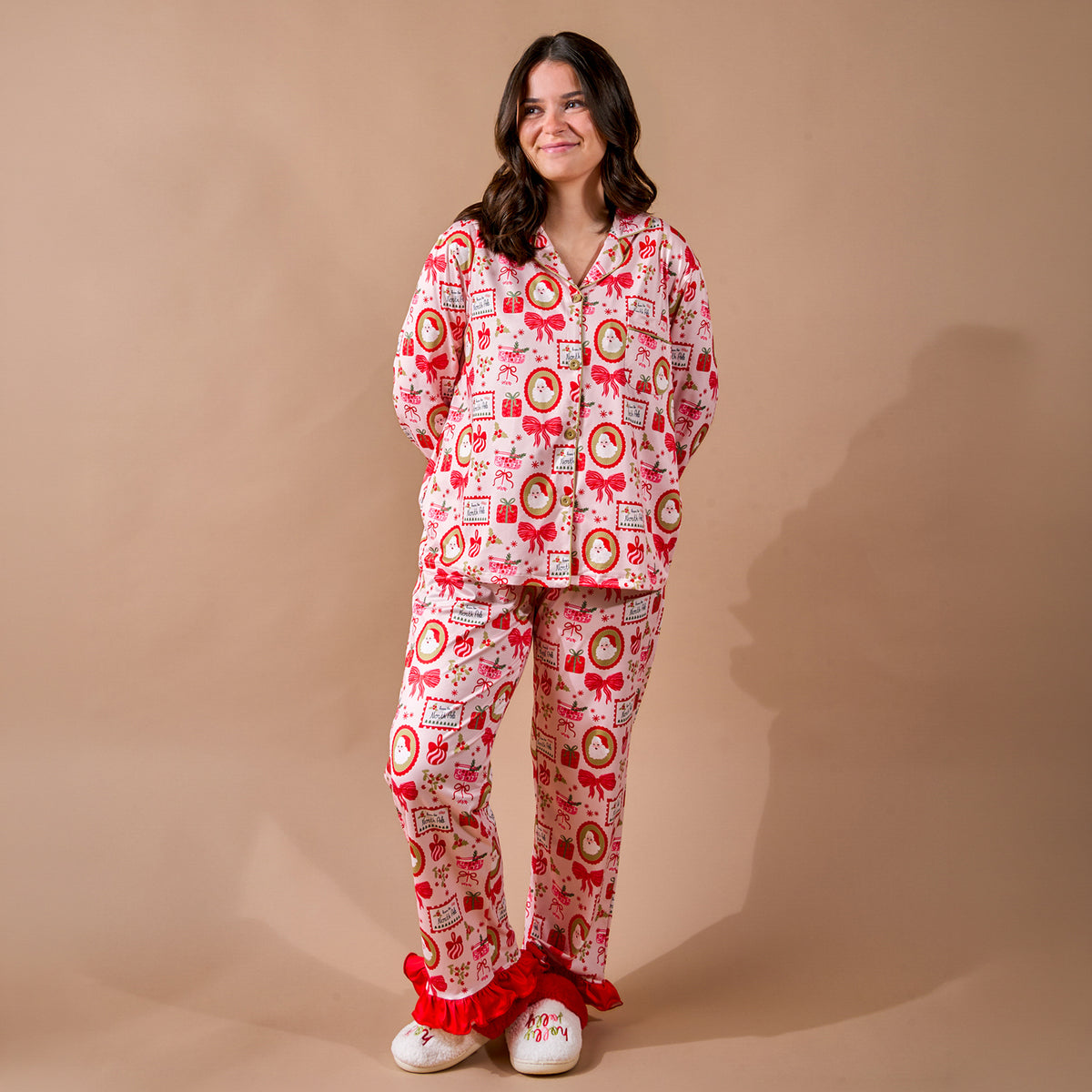A person wearing a Mistletoe Magic Sweet Dreams Pajama Pants Set featuring a long-sleeved button-up top and matching pants with a Christmas-themed print, red ruffles at the ankles, and white slippers.