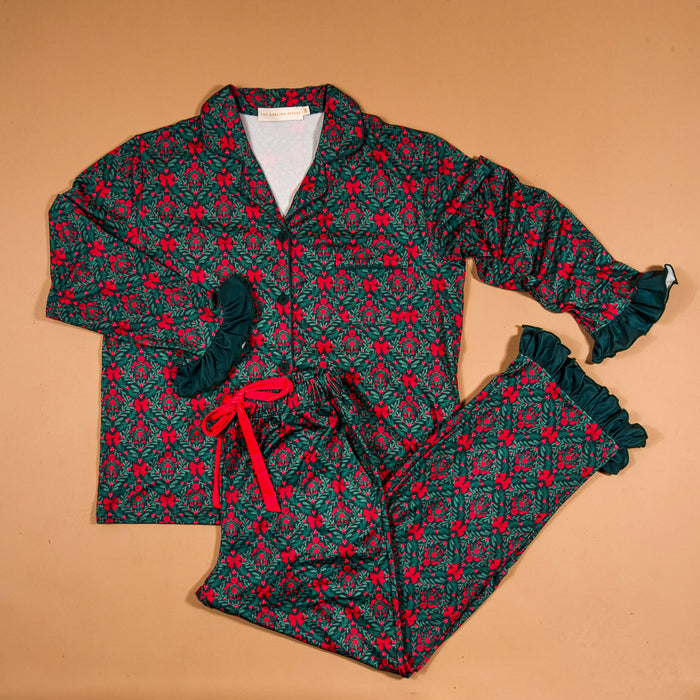 A Merry Moment Sweet Dreams Pajama Pants Set featuring a long-sleeved shirt and matching pants with a red bow and green foliage print, accented by dark green ruffled trim at the cuffs and hem in soft, stretchy fabric.