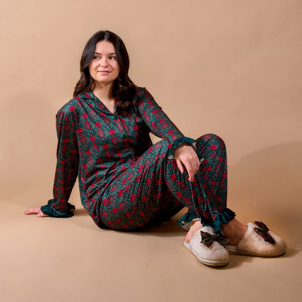 A woman sits on the floor wearing a dark green A Merry Moment Sweet Dreams Pajama Pants Set by The Darling Effect, featuring a red floral print, ruffled cuffs, and fuzzy slippers with bows.