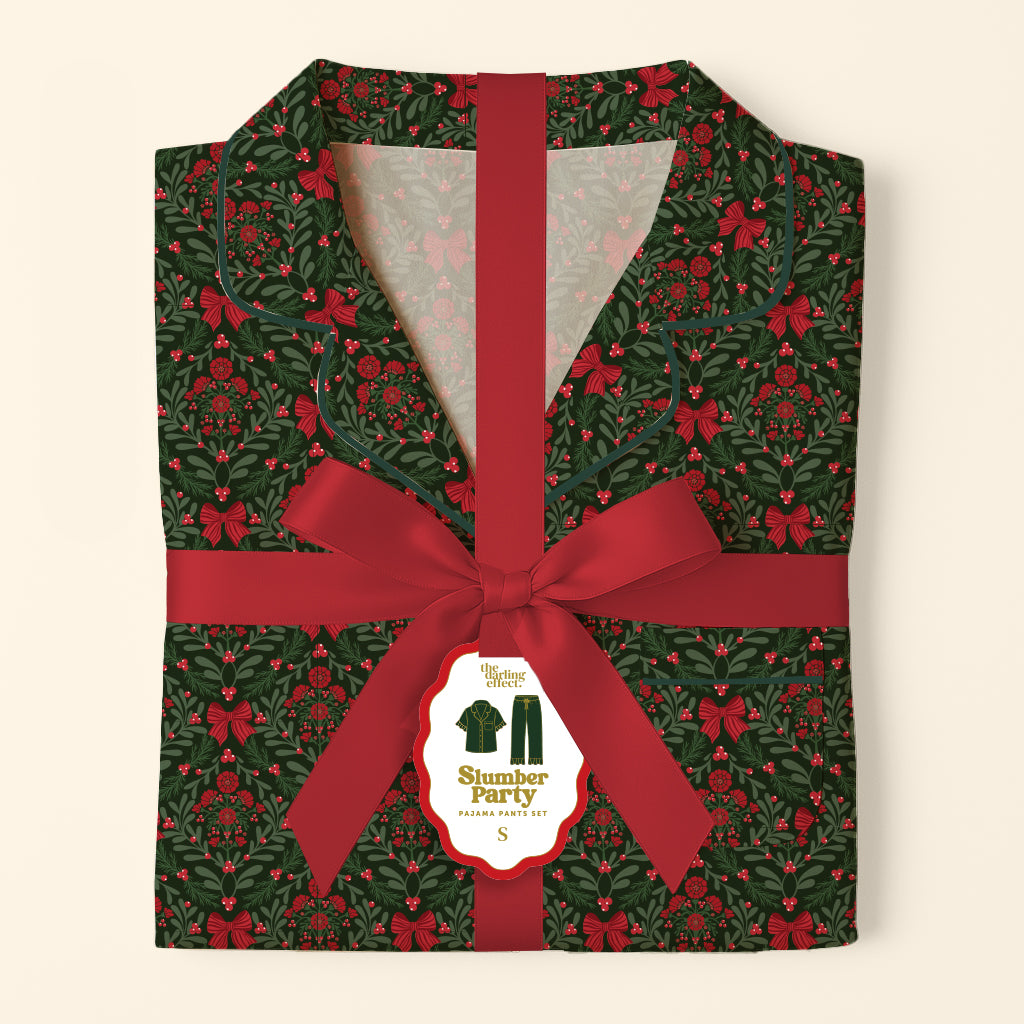 Folded A Merry Moment Sweet Dreams Pajama Pants Set with a dark green pattern of holly, berries, and bows, tied with a red ribbon and a product tag.
