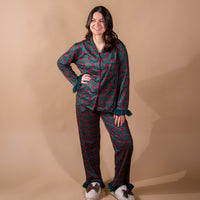 A woman wearing a Merry Moment Sweet Dreams Pajama Pants Set in a red and green holiday pattern, featuring a button-down top and matching pants with ruffled cuffs at the wrists and ankles.