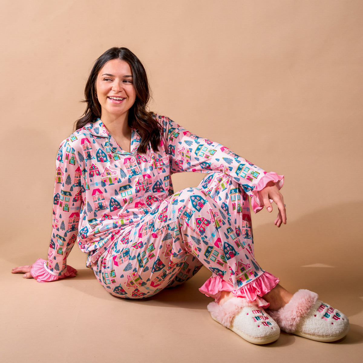 A smiling woman sits on the floor wearing a pink Sugarplum Lane Sweet Dreams Pajama Pants Set by The Darling Effect, featuring a colorful winter village print, ruffled cuffs, and fuzzy white slippers.