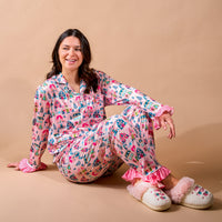 A smiling woman sits on the floor wearing a pink Sugarplum Lane Sweet Dreams Pajama Pants Set by The Darling Effect, featuring a colorful winter village print, ruffled cuffs, and fuzzy white slippers.