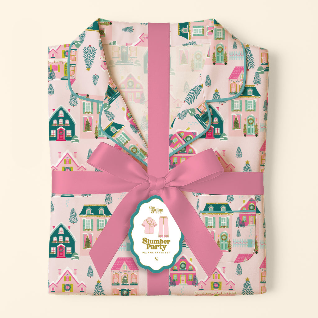 Folded Sugarplum Lane Sweet Dreams Pajama Pants Set with a pink pattern of houses, trees, and snow, tied with a pink ribbon and a product tag.