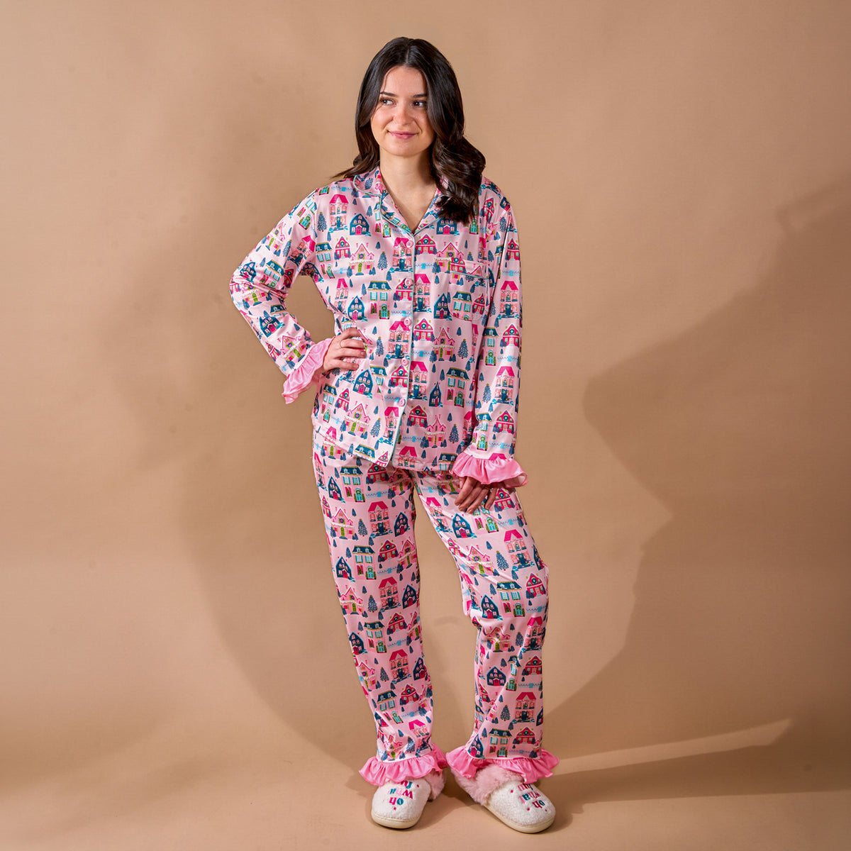 A woman wears a Sugarplum Lane Sweet Dreams Pajama Pants Set by The Darling Effect, featuring a pink long-sleeved button-down top with a village print, ruffled cuffs, matching ruffled-hem pants, and fuzzy slippers.