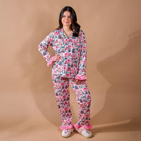 A woman wears a Sugarplum Lane Sweet Dreams Pajama Pants Set by The Darling Effect, featuring a pink long-sleeved button-down top with a village print, ruffled cuffs, matching ruffled-hem pants, and fuzzy slippers.