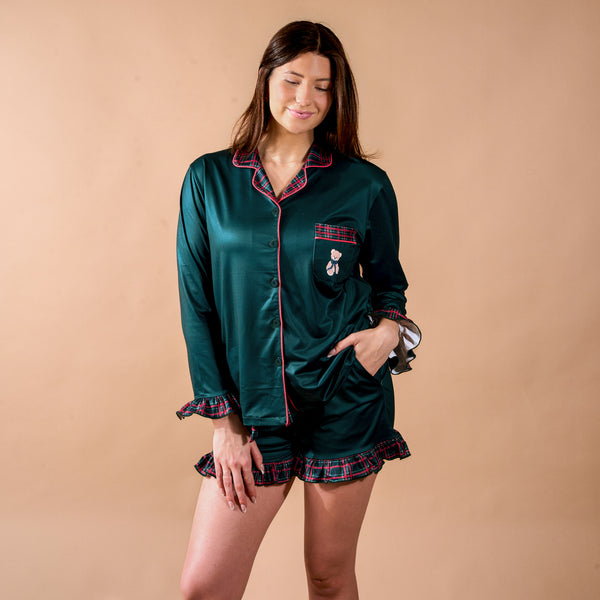 A woman wears the Evergreen Plaid Sweet Dreams Pajama Shorts Set in dark green, featuring a button-down top, shorts with red plaid trim, and a small teddy bear embroidered on the chest pocket.