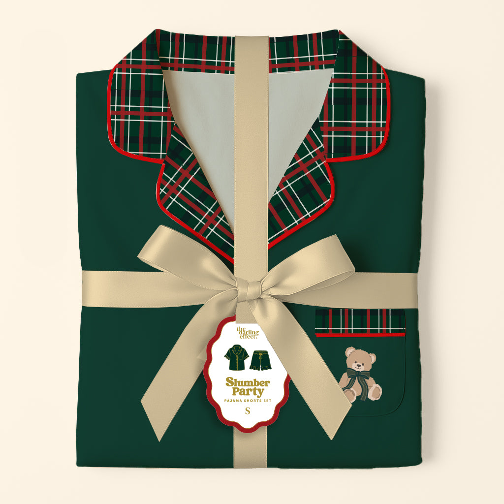 Folded dark green Evergreen Plaid Sweet Dreams Pajama Shorts Set with a red and green plaid collar and pocket, tied with a cream-colored ribbon and a branded gift tag featuring a teddy bear illustration.