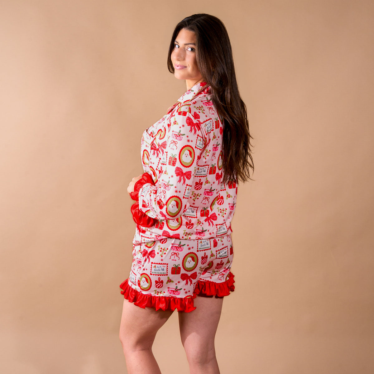 A woman wears the Mistletoe Magic Sweet Dreams Pajama Shorts Set, featuring a white long-sleeve top and matching shorts with a red and green holiday print and red ruffled trim.