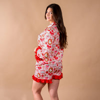 A woman wears the Mistletoe Magic Sweet Dreams Pajama Shorts Set, featuring a white long-sleeve top and matching shorts with a red and green holiday print and red ruffled trim.
