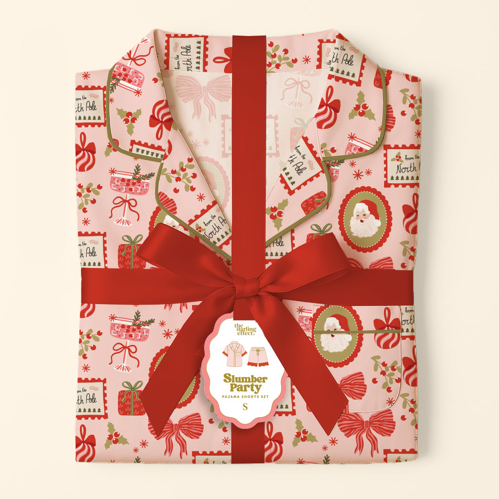 The Mistletoe Magic Sweet Dreams Pajama Shorts Set features pink shorts with a Christmas print of Santa, gift tags, and bows, gift-ready and tied with a red ribbon and a Slumber Party tag.