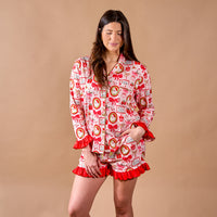 A person wearing The Darling Effect Mistletoe Magic Sweet Dreams Pajama Shorts Set, a cozy long-sleeved top and matching shorts featuring a holiday print of red bows and wreaths with red ruffle trim.