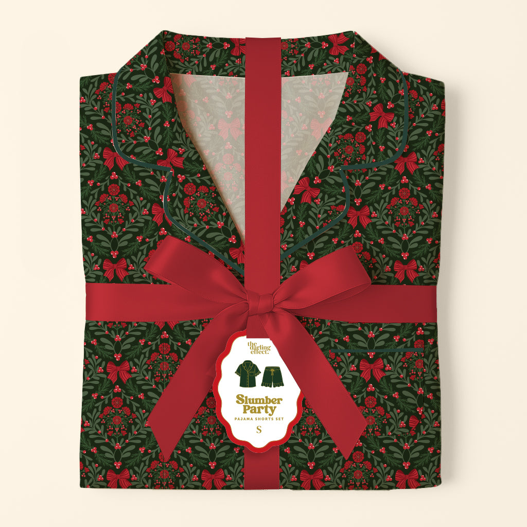 Folded A Merry Moment Sweet Dreams Pajama Shorts Set featuring a dark green Christmas holly and red bow print, wrapped in a red ribbon with a product label.