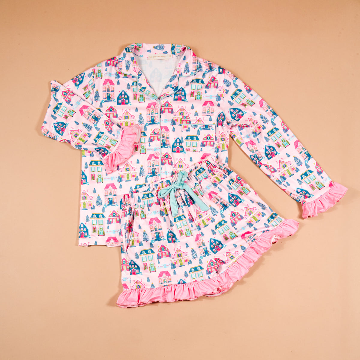 The Darling Effect Sugarplum Lane Sweet Dreams Pajama Shorts Set features a long-sleeved button-down top and matching shorts with a pink, blue, and teal pattern of houses and trees, finished with ruffled sleeve cuffs and hem.