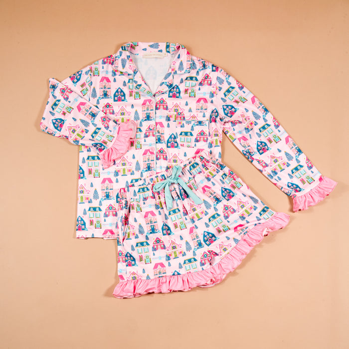 The Darling Effect Sugarplum Lane Sweet Dreams Pajama Shorts Set features a long-sleeved button-down top and matching shorts with a pink, blue, and teal pattern of houses and trees, finished with ruffled sleeve cuffs and hem.