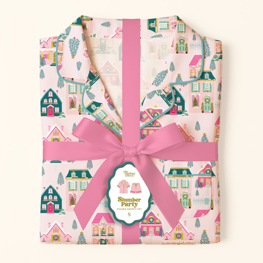 Folded Sugarplum Lane Sweet Dreams Pajama Shorts Set by The Darling Effect featuring shorts with a pink, green, and white holiday house print, wrapped with a pink ribbon and a branded product tag.